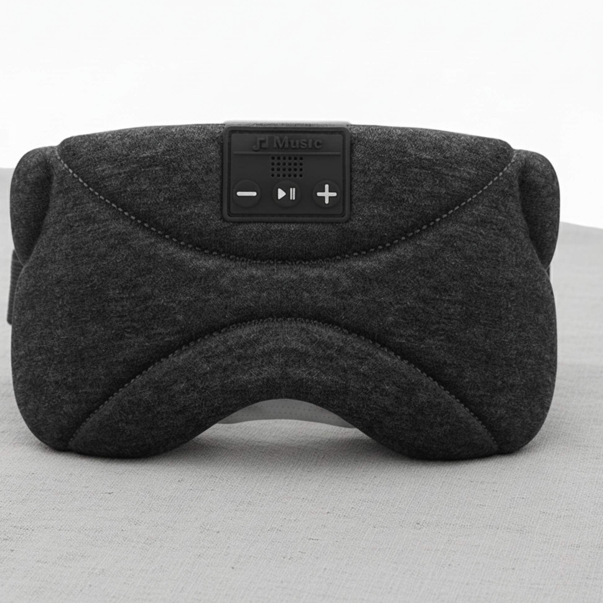 Nærbilde av en mørkegrå Dreamli.no sovemaske med innebygde knapper for avspilling, plassert på et hvitt bord. Bakgrunnen har et lyst, nøytralt trepanel og en grønn plante i en hvit potte. En moderne bordlampe med en bysilhuett på skjermen gir jevn og balansert belysning. Bildet har lav kontrast, myke skygger og en ren, minimalistisk stil som signaliserer ro og søvn.