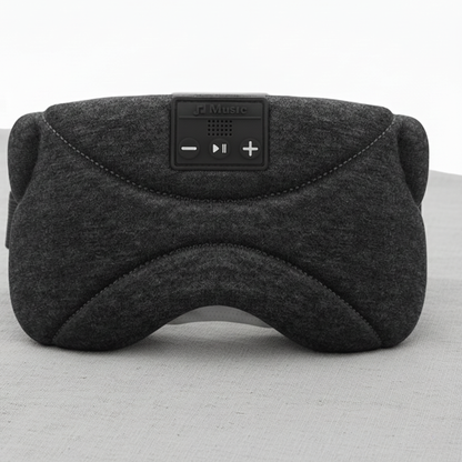 Nærbilde av en mørkegrå Dreamli.no sovemaske med innebygde knapper for avspilling, plassert på et hvitt bord. Bakgrunnen har et lyst, nøytralt trepanel og en grønn plante i en hvit potte. En moderne bordlampe med en bysilhuett på skjermen gir jevn og balansert belysning. Bildet har lav kontrast, myke skygger og en ren, minimalistisk stil som signaliserer ro og søvn.