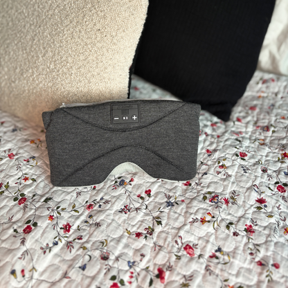 En mørkegrå Dreamli-sovemaske som ligger brettet sammen på en oppredd seng med lyst, hvitt sengetøy, og viser de integrerte knappene for lydkontroll.