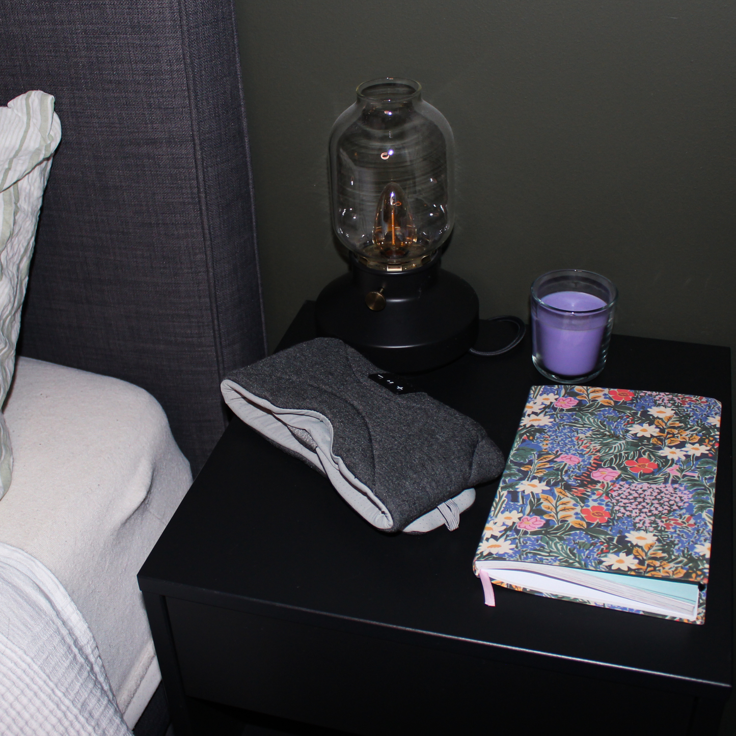 En mørkegrå Dreamli-sovemaske liggende klar på et svart nattbord ved siden av en fargerik notatbok, et tent lilla duftlys og en moderne glasslampe.