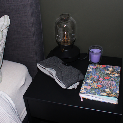 En mørkegrå Dreamli-sovemaske liggende klar på et svart nattbord ved siden av en fargerik notatbok, et tent lilla duftlys og en moderne glasslampe.