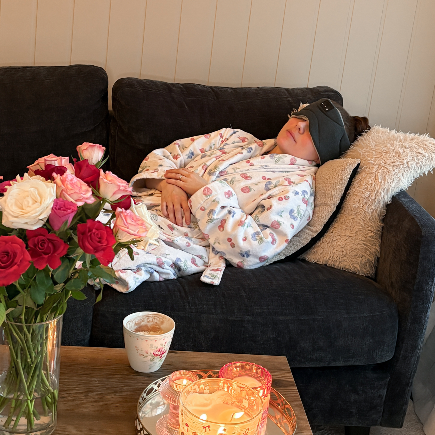 En kvinne som tar en avslappende power nap i en seng med lyst sengetøy, iført en mørkegrå Dreamli sovemaske med synlige Bluetooth-kontroller på siden.